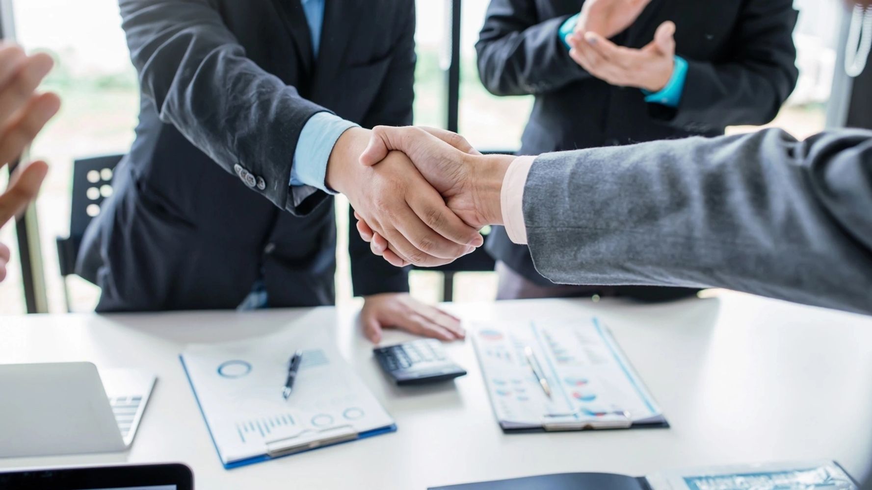 Two professionals in suits shaking hands at a desk with financial documents, a pen, and a calculator